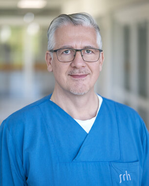 Ein Mann in blauer medizinischer Kleidung steht in einem Flur eines Krankenhauses und schaut direkt in die Kamera.
