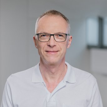 Mann mit Brille und kurzem, grauem Haar, trägt ein weißes Polohemd, lächelt in einem modernen, hellen Raum.