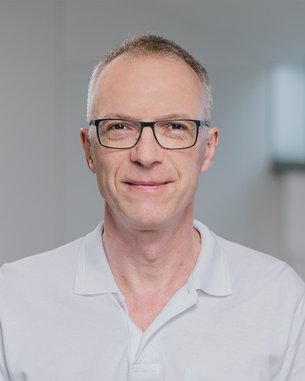 Dr. Straten Ein Mann mit Brille und kurzem, grauem Haar trägt ein weißes Polohemd. Der Hintergrund ist hell und unscharf.