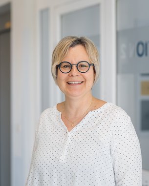 Eine lächelnde Frau mit kurzen, blonden Haaren und runden Brillen steht in einem hellen Raum. Sie trägt ein weißes Oberteil mit Punkten und hat eine goldene Halskette an.
